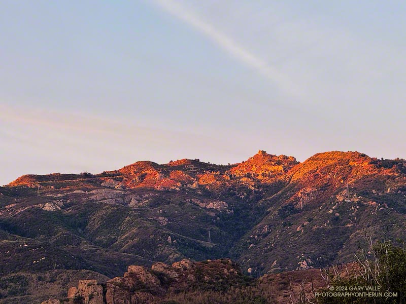The red-tinged light of the rising sun on Castro Crest in the Santa Monica Mountains. The top of Bulldog Mtwy Fire Road is near the skyline in the cetner of the image.